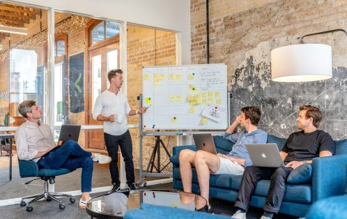 three men sitting while using laptops and watching man beside whiteboard
